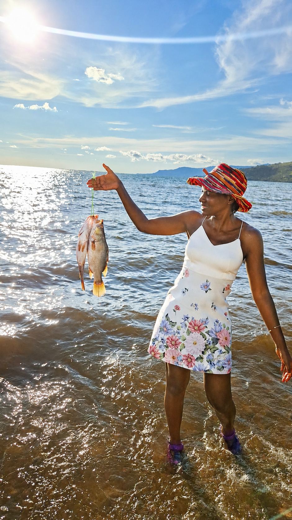Eva standing in Lake Victoria at golden hour, holding a fish, Rusinga Island in the background